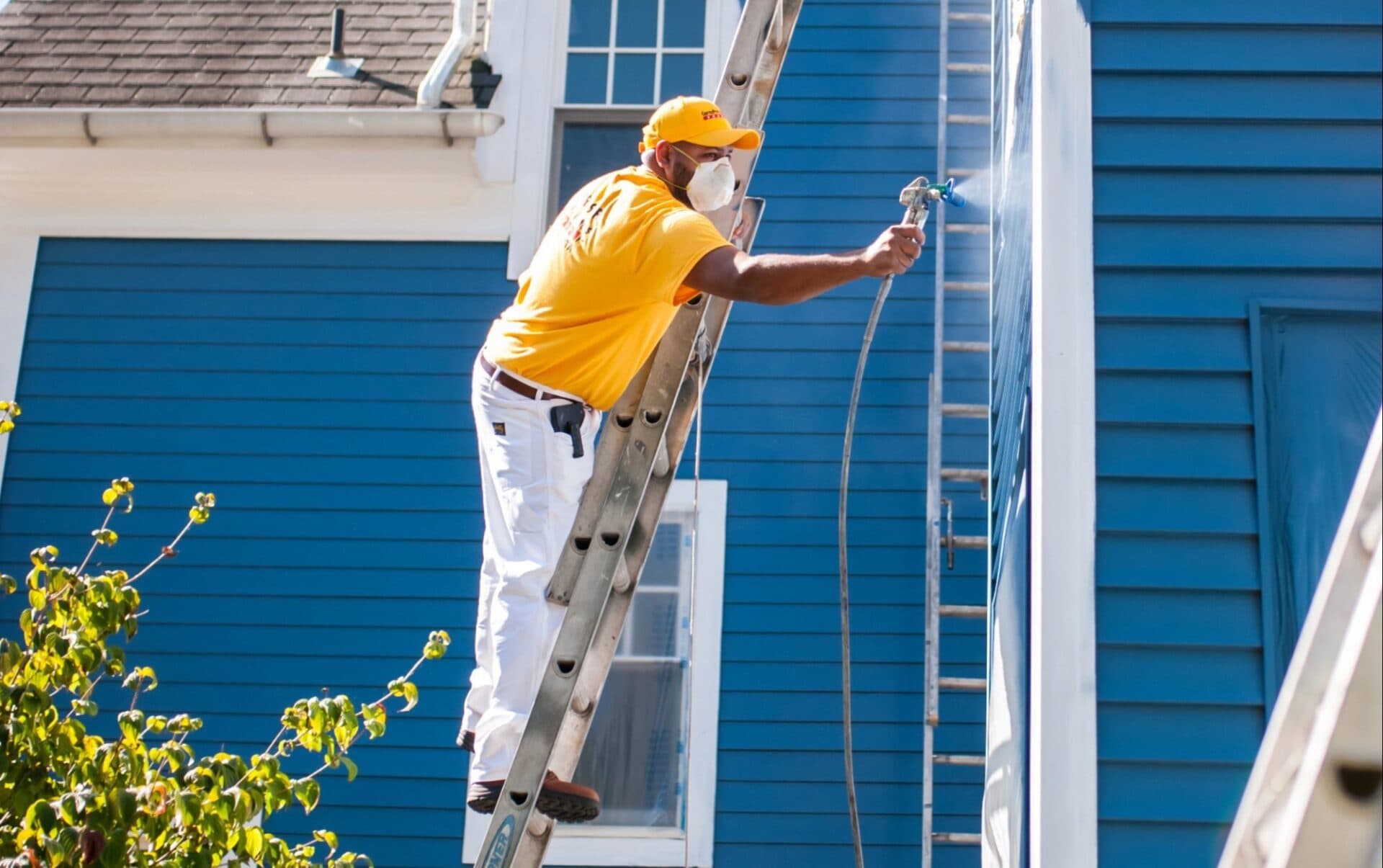 Painter on an extension ladder spray-painting blue siding
