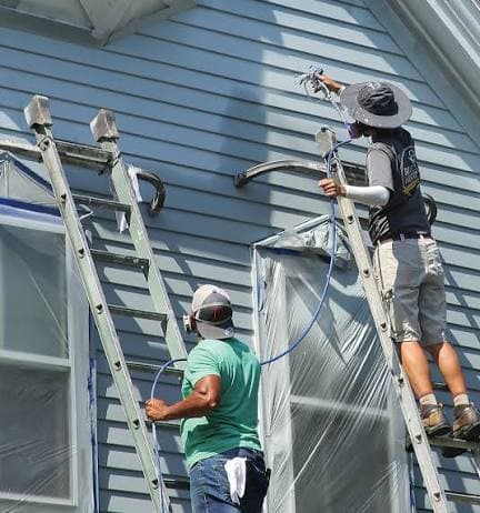 Two E4 crew members prepping and spraying a house exterior from ladders