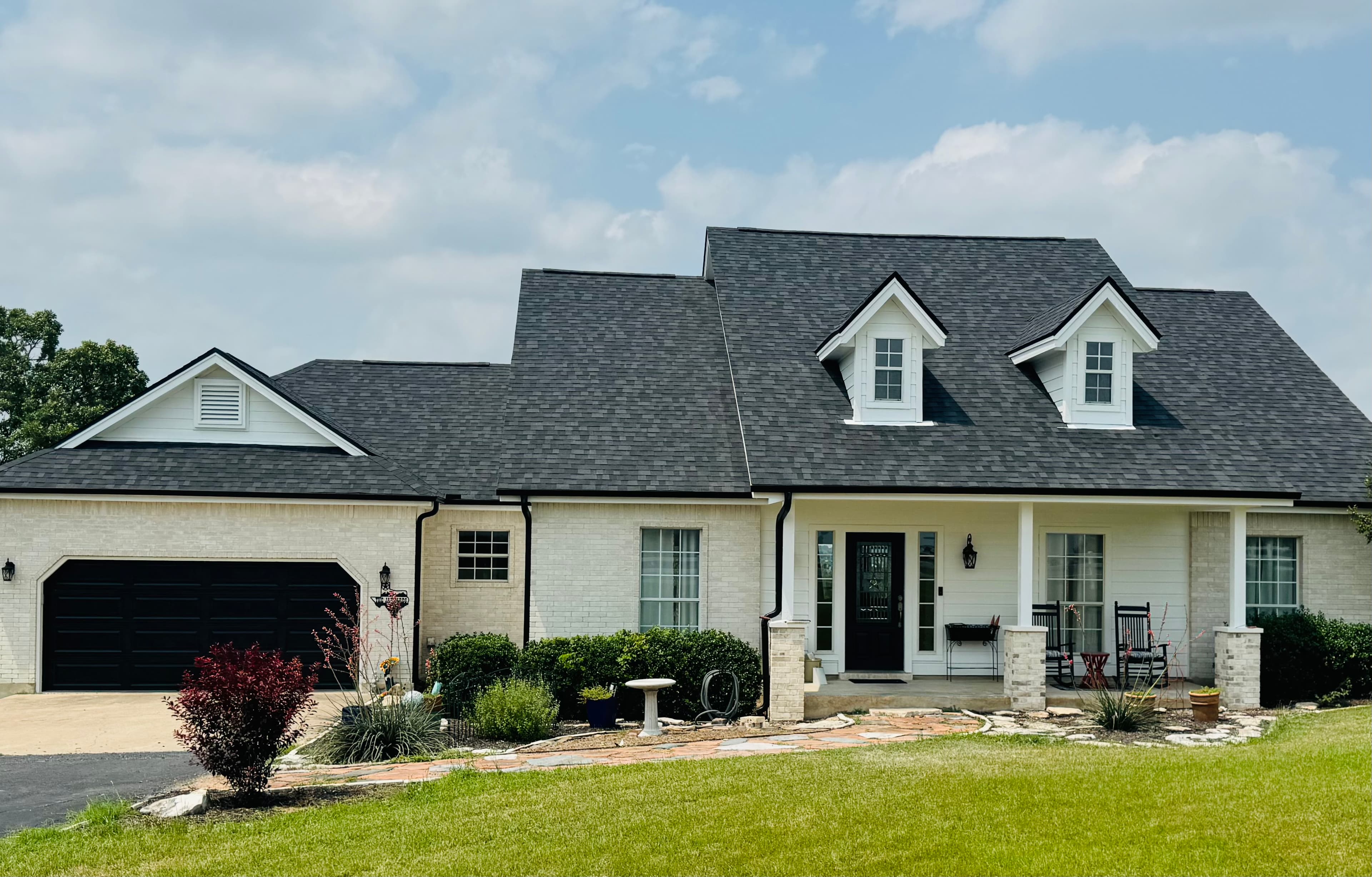 Single-story brick home with a dark grey architectural shingle roof, twin dormers, and a rocking-chair front porch